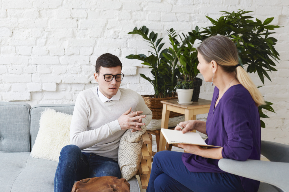 candid-shot-young-man-glasses-talking-about-his-problems-during-psychological-therapy-session-sitting-coach-while-mature-female-psychologist-with-copybook-listening-him-making-notes